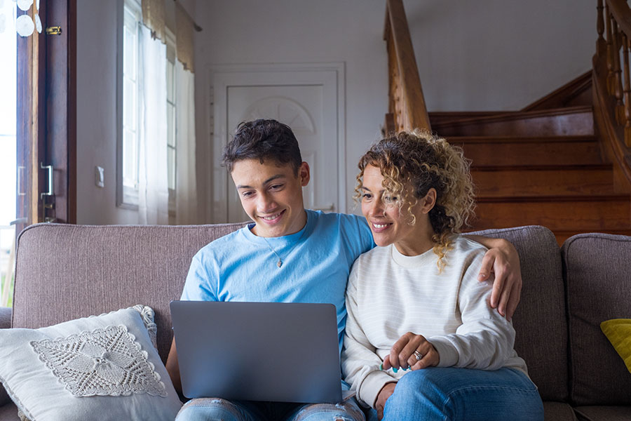 A boy and his mother sitting on the couch, looking at a laptop and smiling.