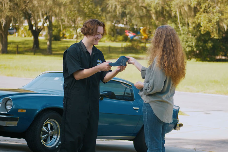 A mother handing her teenage J-Tech student a branded J-Tech hat and sending him off to school in his antique muscle car.