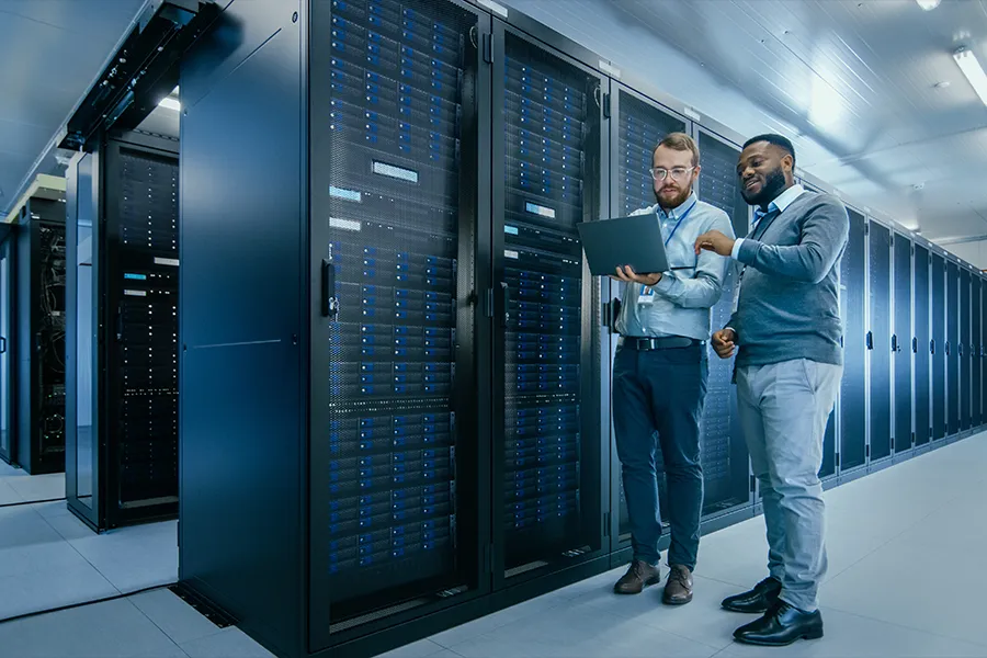 Two system administrators standing in a server room beside a rack, holding a laptop and diagnosing an issue.